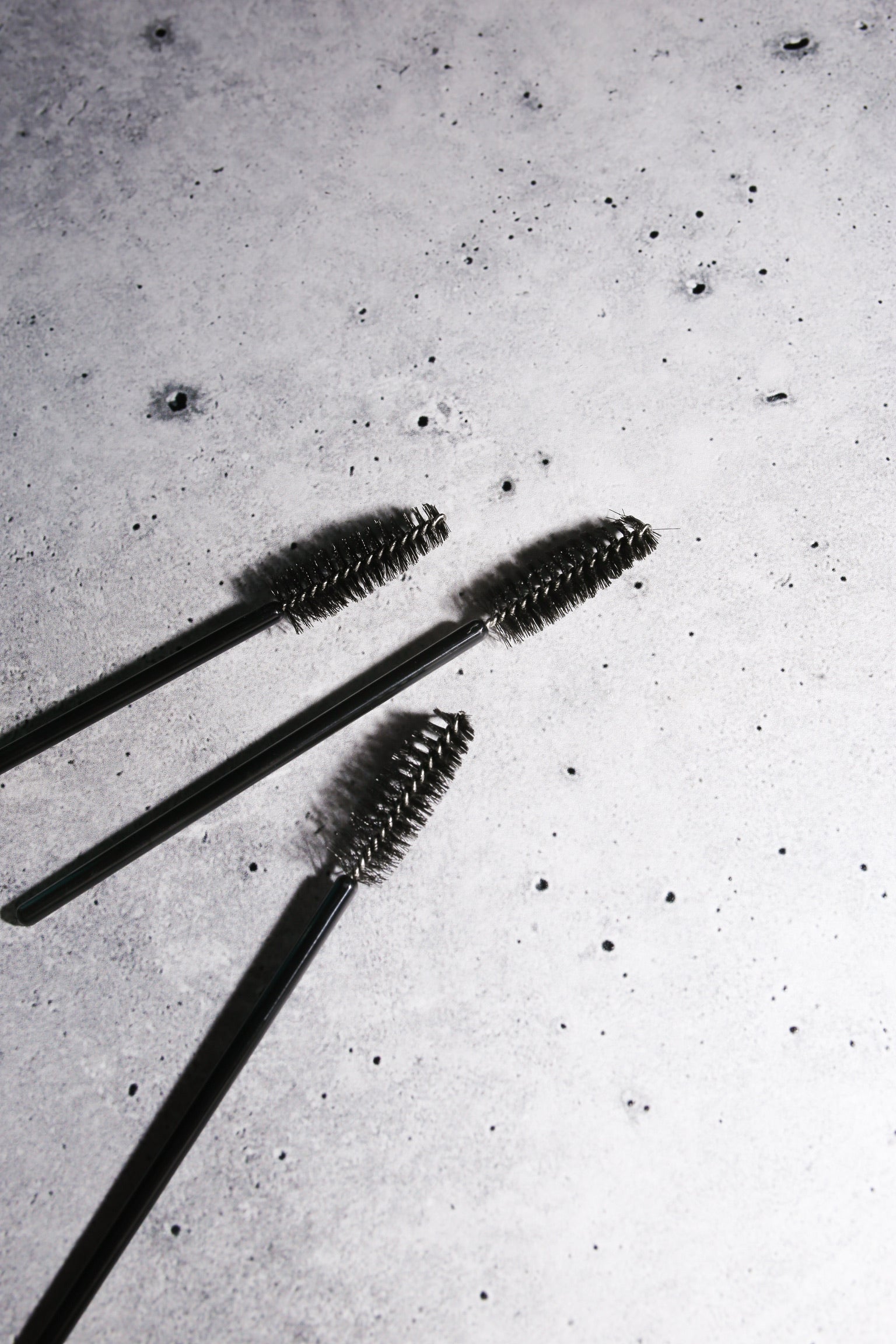Three black mascara brushes on a textured gray surface